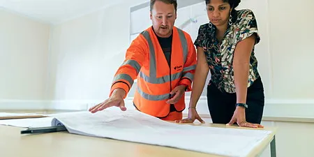 Reflektörlü yelekli çalışan ve iş arkadaşı masada teknik çizim inceliyor